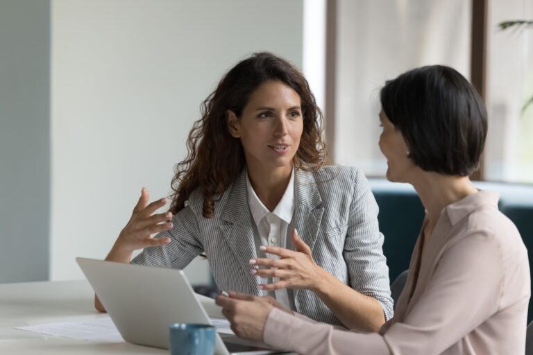 Two,Business,Women,Sit,At,Desk,Discuss,Project,Details,,Diverse